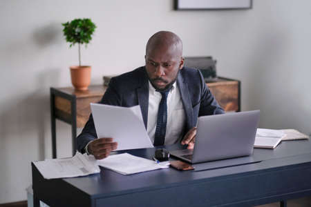 Young African executive reading documents and working on a laptop while sitting at his desk in an office. soft focusの写真素材