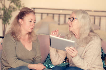 Senior woman retiree showing good news on digital computer tablet to grown daughter, relaxing together on couch indoors, different generations hobby pastime.の写真素材