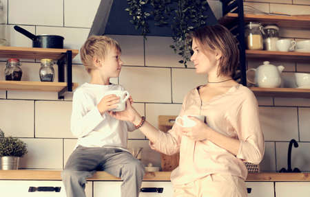 young mom and son look at each other. Happy family in the morning. Beautiful woman with her son having breakfast in kitchen together, son looking at mother and smilingの写真素材