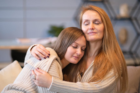 Cheerful young woman hugging aged mother sitting on sofa while resting together in living room at...