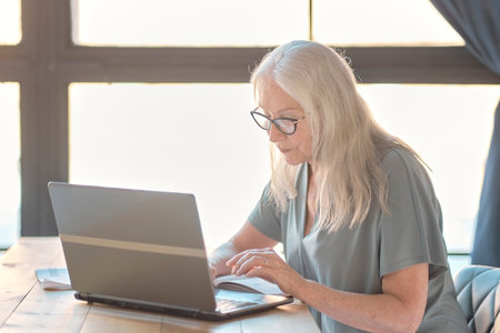 Senior woman using laptop for websurfing in her kitchen. The concept of senior employment, social security. Mature lady sitting at work typing a notebook computer in an home office.の写真素材