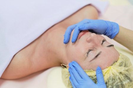 Close up portrait of charming woman with closed eyes enjoying skincare procedure. Masseuse arms touching client chin and nose. Anti-aging effectの写真素材