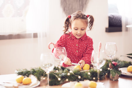 Cute curly toddler girl standing at a Christmas dinner table settling the dishes preparing to celebrate Xmas Eve a decorated dining room with tree and lightsの写真素材