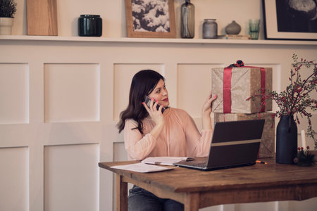 Pretty young lady sitting at desk and typing on laptop and talking on the phone with present boxes icons. box receive good online shop purchase at home, post mail shipping delivery conceptの写真素材