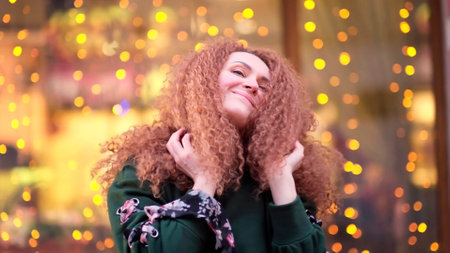 cheerful woman is touching her curly red hair, fooling around against the background of glowing bulbs.の写真素材