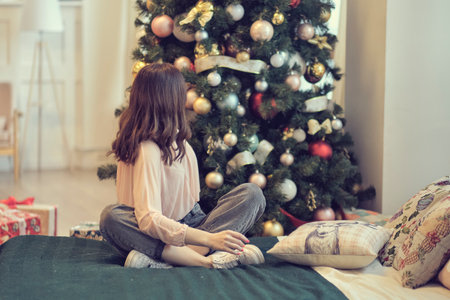 young woman sits on a bed against the background of a Christmas tree. concept of starting life anew after celebrations.の写真素材