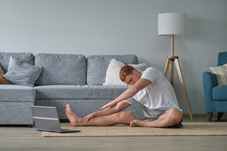 oung cheerful man doing morning gymnastic at home, sitting on yoga mat and stretching, copy space....