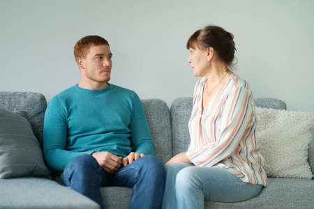 elderly mother tells her son her news. Cheerful elderly woman sitting on the sofa next to his adult son. relations between different generations.の写真素材