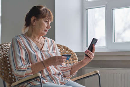 Portrait senior woman shopping online or pays utility bills through the personal account of the online bank, using credit card and smartphone at home.の写真素材