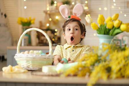 child boy wearing bunny ears sitting in the kitchen at the table. child holding painted eggs preparing for easter. funny surprised childの写真素材