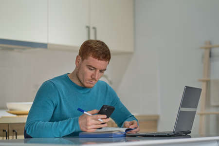 Entrepreneur man on the phone working with a laptop in the kitchen at home. freelancer is negotiating on a smartphoneの写真素材