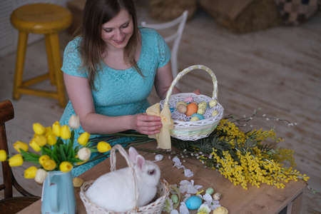 young woman decorates Easter basket in cozy kitchen interior. table kitchen with flowers, easter colorful eggs, presents, pet white rabbit sitting in a basket.の写真素材