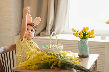 child boy wearing bunny ears sitting in the kitchen at the table. child holding painted eggs preparing for easter. funny boy showing thumbs upの写真素材