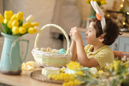 child boy wearing bunny ears sitting in the kitchen at the table. child holding painted eggs preparing for easter. funny boy showing thumbs upの写真素材