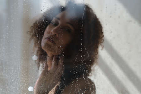 Sensual portrait of young woman taking a shower. Defocused female looks through the glass of the shower stall.の写真素材