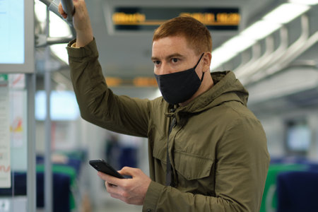 Handsome guy in respirator use a smartphone and keep social distancing to crowd while commuting in the metro or train. Infection and air pollution prevention conceptの写真素材