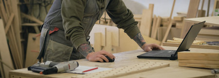 Concentrated adult craftsman checking information on laptop while working in carpentry workshop. profession, carpentry, technology and people conceptの写真素材