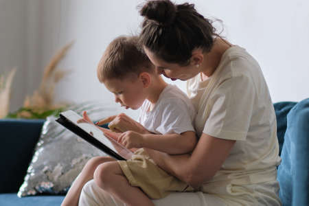 mother and son with tablet at home. Mother showing media content on line to her son in a tablet sitting on a couch in the living room in a house interiorの写真素材