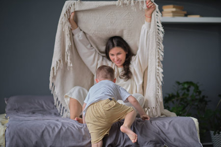 mom and child have fun in the bedroom on the bed, play under the blanket. Young mother with her little son dressed playing in the bed at the weekend together, lazy morning, warm and cozy scene.の写真素材