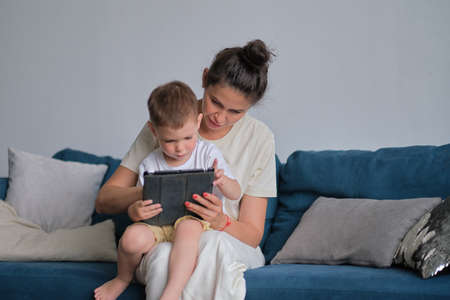 mother and son with tablet at home. Mother showing media content on line to her son in a tablet sitting on a couch in the living room in a house interiorの写真素材