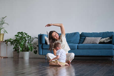 Happy mindful single mother doing morning exercises in yoga pose with kid playing at home. morning exercises in pajamas. child interferes with the workoutの写真素材