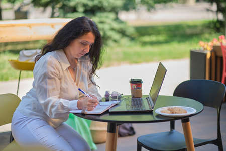Mature woman at coffee shop. Portrait of confident mature professional woman sitting on summer terrace in cafe, using laptop computer for work, laughing happily indoors.の写真素材