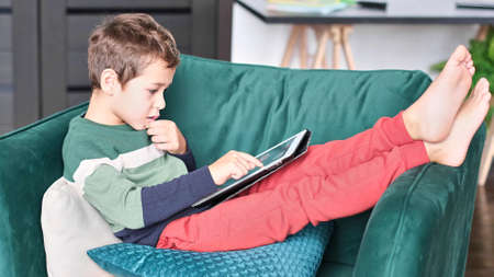 Cheerful little boy smiling while sitting on couch and using tablet at home. Modern kid and education technology. little boy looking at touch pad at home.の写真素材