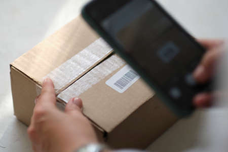 Small business owner standing at her shop arranges delivery through the application on a smartphone. online store seller entrepreneur packing package post shipping box preparing delivery parcel.の写真素材