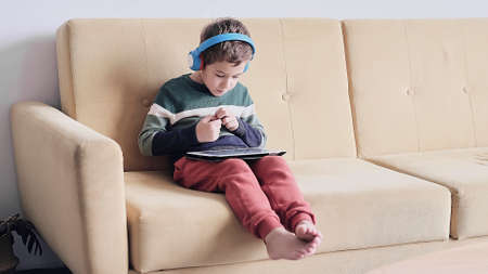 Cheerful little boy smiling while sitting on couch and using tablet at home. Modern kid and education technology. little boy looking at touch pad at home.の写真素材
