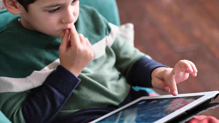 Cheerful little boy smiling while sitting on couch and using tablet at home. Modern kid and education technology. little boy looking at touch pad at home.の写真素材