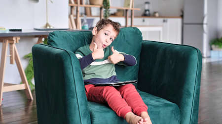 Cheerful little boy smiling while sitting on couch and using tablet at home. Modern kid and education technology. little boy looking at touch pad at home.の写真素材