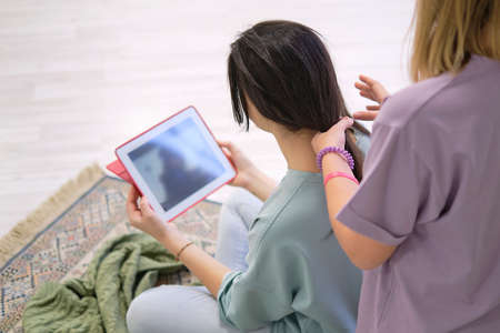 girl makes a braid to her friend. Hair weaving hairstyles. Girlfriend braids her hands with ringlets. Two young multinational girls spending time together, pajama party.の写真素材