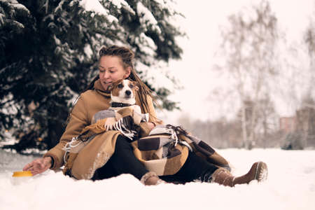 playful woman with dog. stylish hipster woman hugging and smiling cute puppy in snowy cold winter park. moments of true happiness.の写真素材