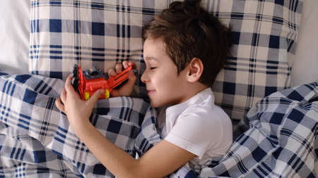 cheerful funny boy lies in bed and hugs his favorite toy. The boy wakes up with a cheerful mood. little kid boy sleeping in bed with toy.の写真素材