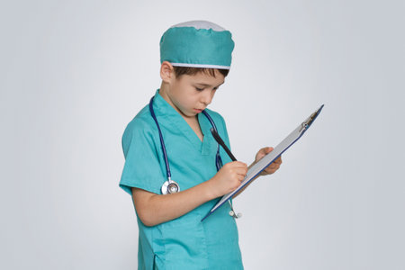 little boy in medical uniform character holds clipboard and writing diagnosis. serious swarthy boy writes down a treatment plan on clip-on tablet, isolated on whiteの写真素材