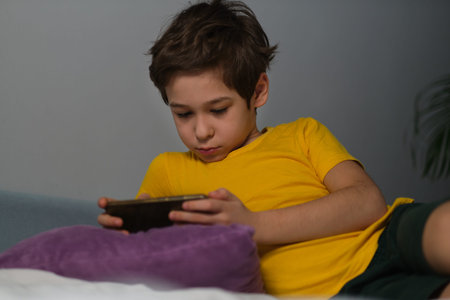 Little asian boy playing on the phone and chewing on his feet, the concept of stress in a child,...