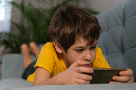 boy with smartphone or game console in hands, playing or engaging over social media while lying on a...