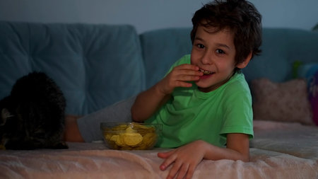 boy eats chips and watches TV at home with curiosity. Leisure time for children watching TV. Fast...