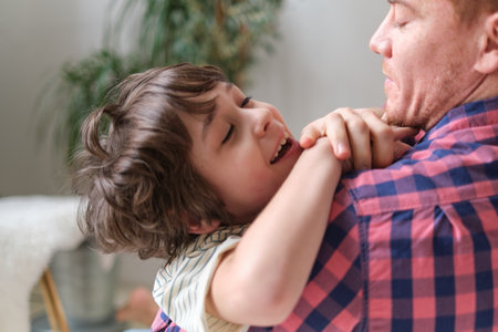 Father and son embracing with joy, sitting on a living room couch, smiling at the camera. The Importance of Family Bonding and Quality Time. Father-Son Relationships: Building Strong Foundationsの写真素材