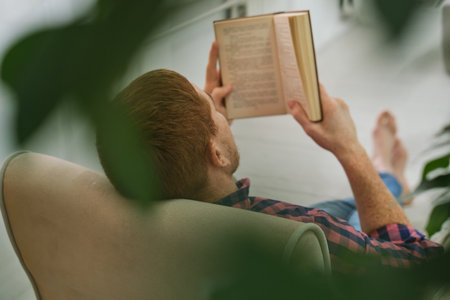 Literary Oasis. Man taking a breather with a good read, while green plants create a soothing indoor sanctuary. Homebound Tranquility.の写真素材