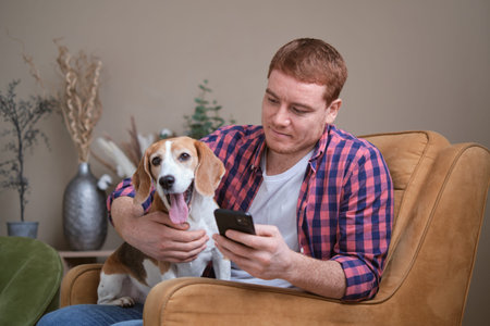 Man and his loyal Beagle spending a lazy afternoon on the sofa: browsing social media and petting his furry companion for some much-needed relaxation.の写真素材