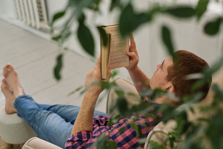Man leisurely reading a book in a comfortable chair, nestled within verdant foliage indoors. Natures Reading Nook.の写真素材