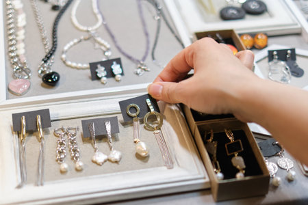 womans hand delicately selects a handmade earring from a display, showcasing an array of artisanal jewelry in the background. supporting local artisans, and unique fashion accessoriesの写真素材