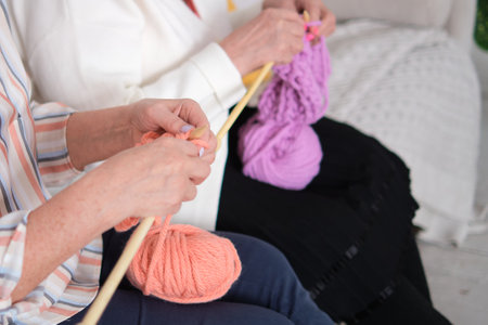 tender moment of two elderly women knitting with wooden needles and pink yarn, illustrating the age-old tradition of sharing stories, knowledge, and companionship through the art of crafting.の写真素材