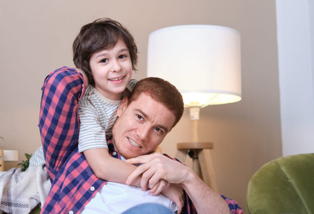 Father and son share a heartwarming embrace on a comfortable sofa, their eyes filled with love and...