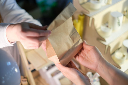 female vendor at a bustling fair passes a paper bag with purchases to a shopper, illustrating the...