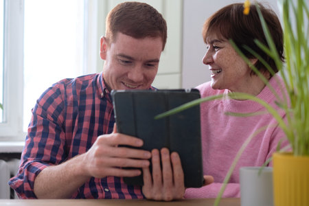 mother and her adult son sitting at a table, sharing knowledge as he teaches her how to navigate a tablet. help seniors stay connected in a digital worldの写真素材