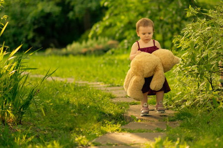 Little toddler girl holding a large teddy bear walks along a sunlit path in a parkの写真素材