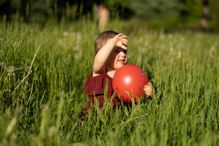 young child lost in the world of a red ball, sitting in the summer grass, under a radiant sun. Kids sunscreen product , early education toys cataloguesの写真素材