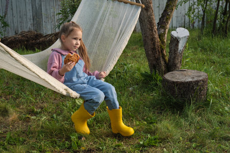 As roots connect with the soil, so does a little girl with her surroundings. In a hammock, she eats a bun, portraying the essence of nourishment, both physical and emotional, during formative yearsの写真素材
